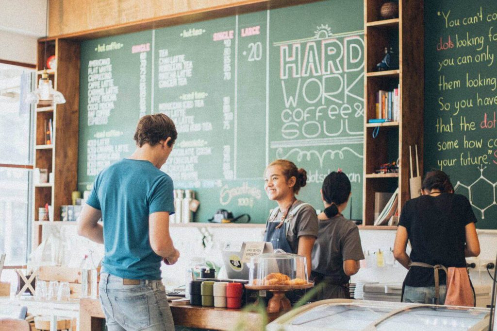 مشتری در حال سفارش در کافه چی با دکوراسیون مدرن و منوی نوشته شده روی تخته